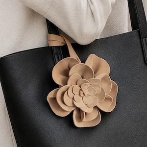 May include: A black tote bag with two shoulder straps and a large, layered beige flower charm attached to one of the handles. The bag has a smooth, matte texture and a structured rectangular shape, providing a contrast to the soft, light-colored coat.