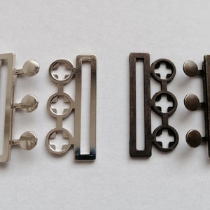 May include: Two metal belt buckles, one silver and one bronze. Each buckle has three round holes and a rectangular frame.