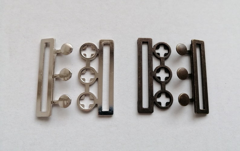 May include: Two metal belt buckles, one silver and one bronze. Each buckle has three round holes and a rectangular frame.