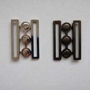 May include: Two metal belt buckles with three round decorative accents. The buckle on the left is silver and the buckle on the right is black.