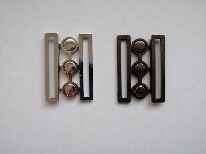 May include: Two metal belt buckles with three round decorative accents. The buckle on the left is silver and the buckle on the right is black.