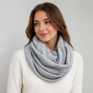 May include: A light gray infinity scarf with a soft, textured knit fabric. The scarf is wrapped in multiple loops around the neck, creating a voluminous and layered appearance. The material has a heathered look with subtle variations in tone.