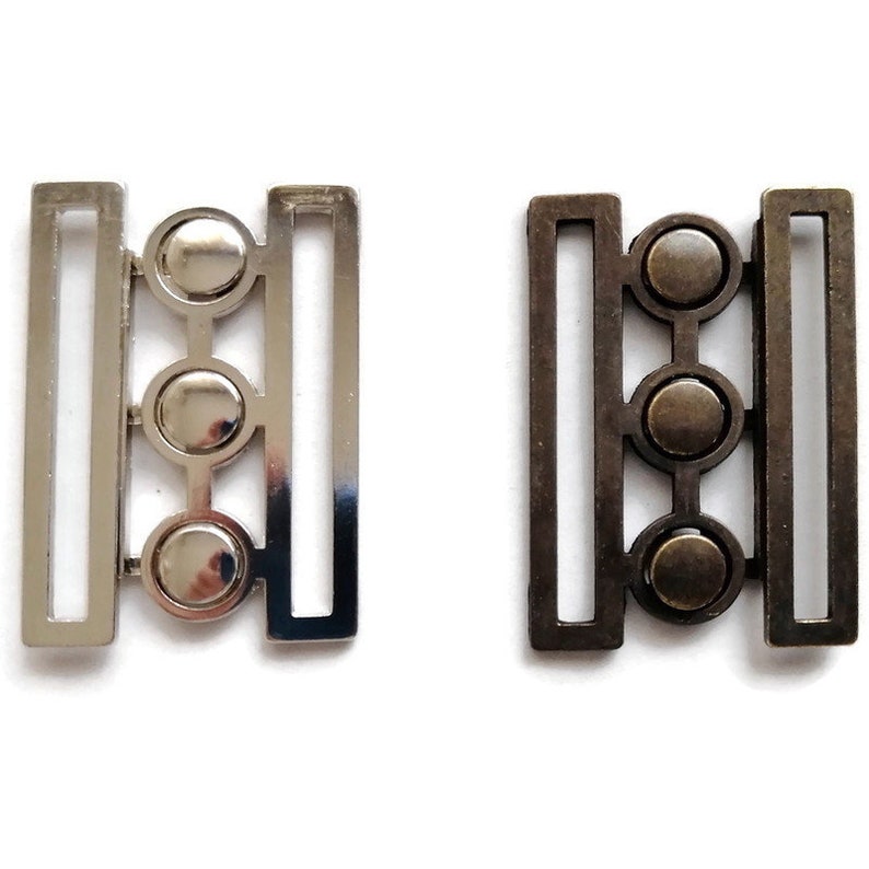 May include: Two metal belt buckles with three round decorative elements. The buckle on the left is silver and the buckle on the right is bronze.