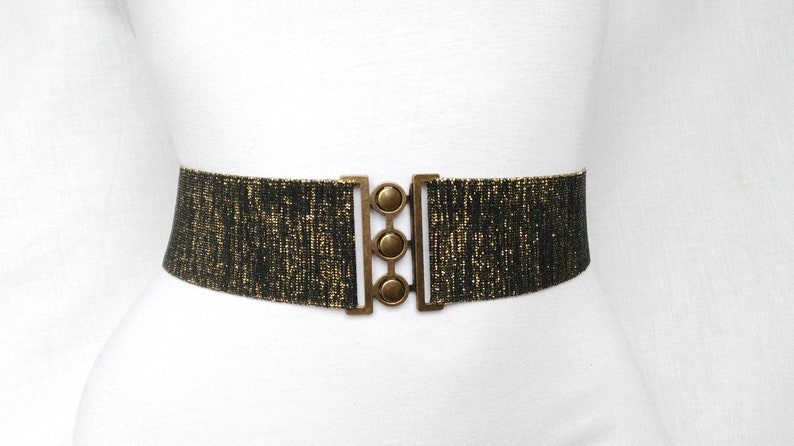 May include: A black and gold elastic belt with a gold metal buckle. The buckle has three round decorative pieces.