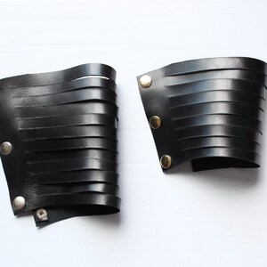 May include: Two black leather wrist cuffs with horizontal cut-out strips. Each cuff has metal snap closures. The cuffs are displayed on a white background. The cuffs are likely for cosplay or fashion.