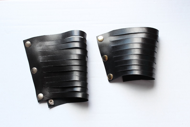 May include: Two black leather wrist cuffs with horizontal cut-out strips. Each cuff has metal snap closures. The cuffs are displayed on a white background. The cuffs are likely for cosplay or fashion.