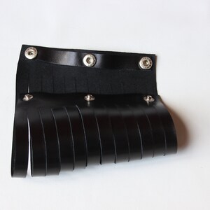 May include: Black leather wrist cuff with fringe detail. The cuff features a strap with three snap closures and multiple vertical leather strips hanging down. The accessory is designed to be worn around the wrist.