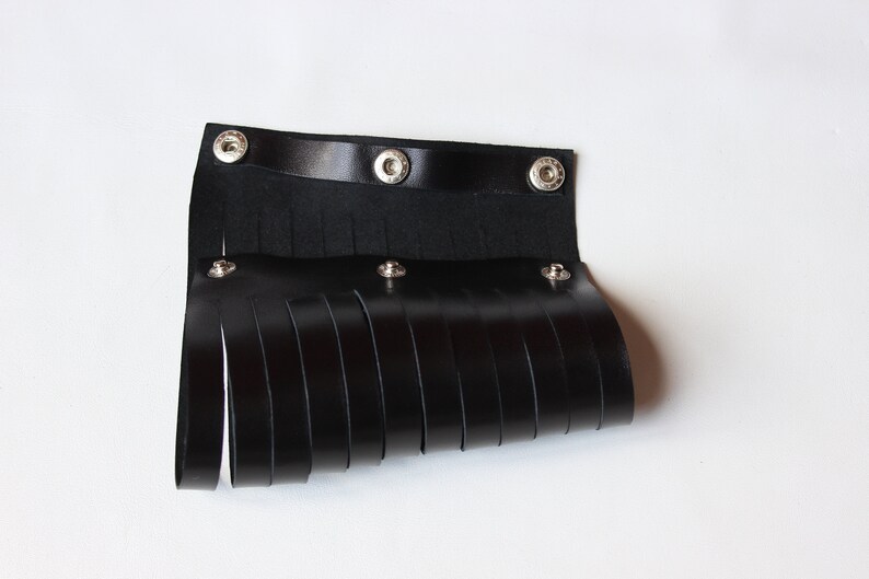 May include: Black leather wrist cuff with fringe detail. The cuff features a strap with three snap closures and multiple vertical leather strips hanging down. The accessory is designed to be worn around the wrist.