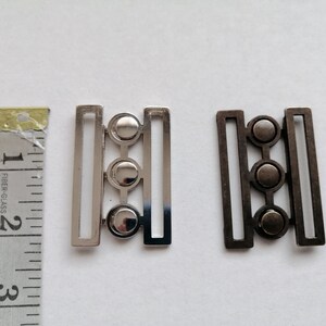 May include: Two metal clasps with three round decorative elements. The clasp on the left is silver and the clasp on the right is black.