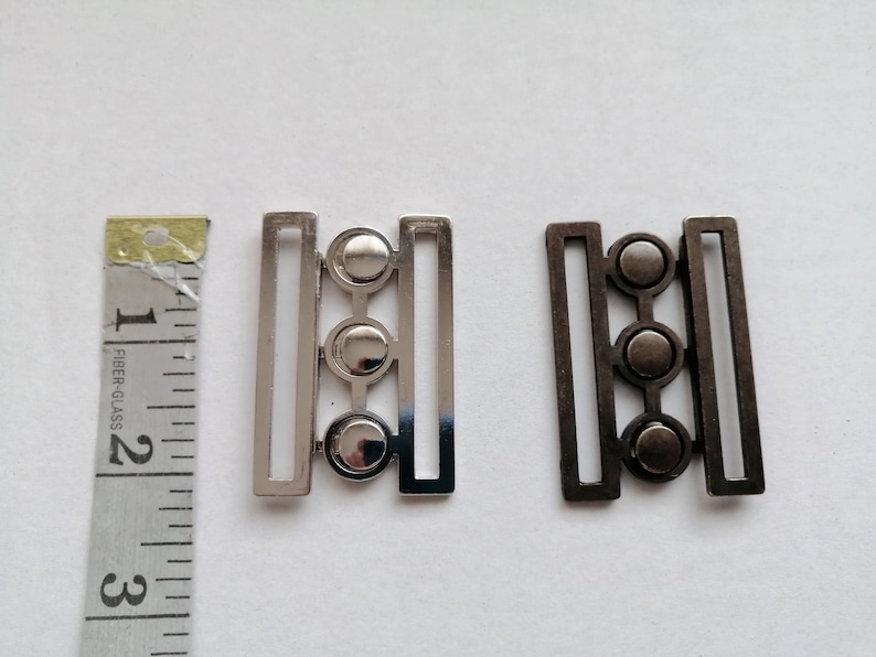 May include: Two metal clasps with three round decorative elements. The clasp on the left is silver and the clasp on the right is black.