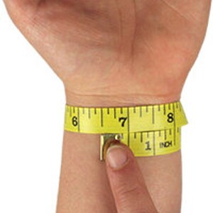 May include: A yellow measuring tape wrapped around a wrist, showing a measurement of approximately 7 inches. The tape is marked with black numbers and inch increments, with the word "INCH" visible.