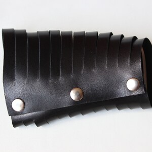 May include: Black leather wrist cuff with a series of vertical, fringed strips. The cuff is secured with three silver-colored snap closures. The design is a bold statement piece.