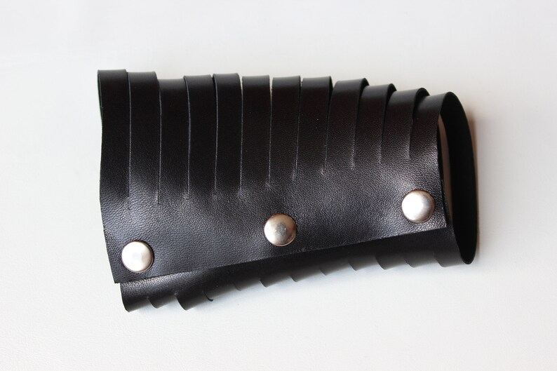 May include: Black leather wrist cuff with a series of vertical, fringed strips. The cuff is secured with three silver-colored snap closures. The design is a bold statement piece.