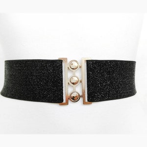 May include: A black elastic belt with a silver buckle featuring three round accents. The belt is adorned with a subtle sparkle.