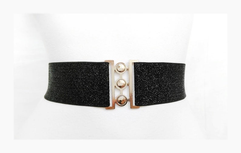 May include: A black elastic belt with a silver buckle featuring three round accents. The belt is adorned with a subtle sparkle.