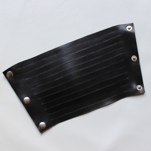 May include: A black leather wrist cuff with horizontal stitched detailing. The cuff is rectangular with rounded corners and silver snap closures. The cuff is shown on a white background.