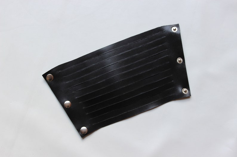 May include: A black leather wrist cuff with horizontal stitched detailing. The cuff is rectangular with rounded corners and silver snap closures. The cuff is shown on a white background.