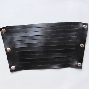 May include: Black leather wrist cuff with horizontal cut-out slits. The cuff is secured with three silver-colored snaps on each end. The leather has a smooth texture and is set against a white background.