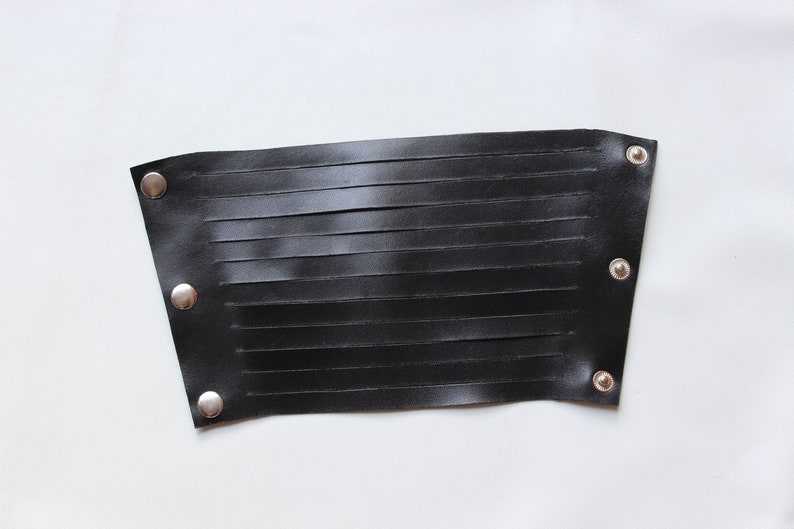 May include: Black leather wrist cuff with horizontal cut-out slits. The cuff is secured with three silver-colored snaps on each end. The leather has a smooth texture and is set against a white background.