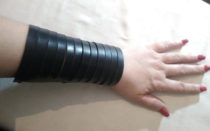 May include: Black leather wrist cuff with multiple horizontal straps. The cuff is secured with small silver-colored rivets. The image shows the cuff worn on a person's wrist, with the hand and fingers visible.
