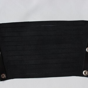 May include: Black leather wrist cuff with horizontal cut-out lines. The cuff has a rectangular shape with rounded corners and is secured with silver snap closures. The cuff is shown against a white background.