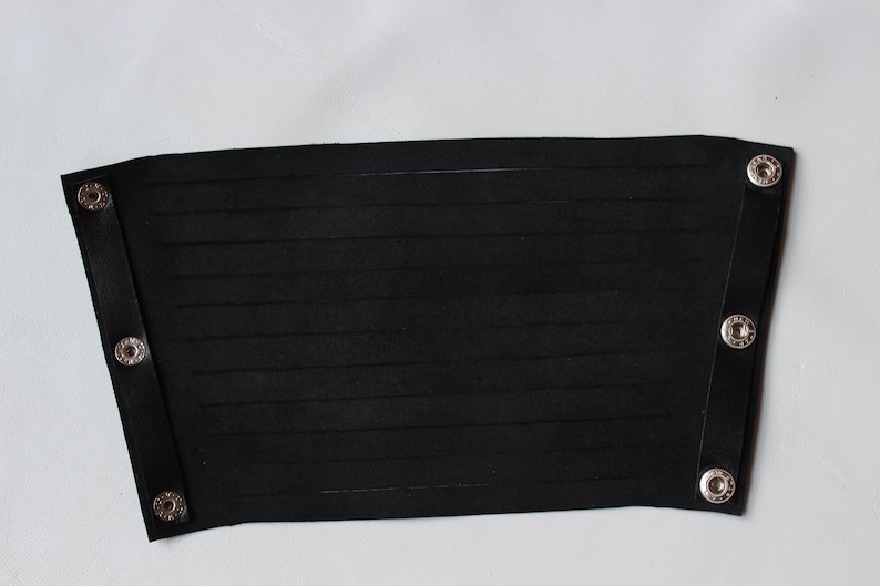 May include: Black leather wrist cuff with horizontal cut-out lines. The cuff has a rectangular shape with rounded corners and is secured with silver snap closures. The cuff is shown against a white background.