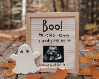 May include: A wooden sign with the text "Boo! We've been keeping a spooky little secret brewing until Oct 2025."  A white ghost figurine is to the left of the sign.  The sign is on a wooden surface with fall leaves.