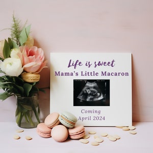 Könnte beinhalten: Ein weißer gerahmter Schild mit dem Text "Life is sweet Mama's Little Macaron" und einem Schwarzweiß-Ultraschallbild. Das Schild sagt auch "Coming April 2024".  Rosa und weiße Blumen in einer Glasvase und pastellfarbene Macarons befinden sich auf einer rosa Oberfläche.