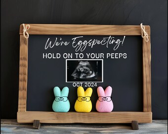 May include: A wooden chalkboard with the text "We're Eggspecting! Hold on to your peeps" and an ultrasound image. The date "OCT 2024" is written below the image. Three pastel-colored bunny-shaped candies are in front of the chalkboard.