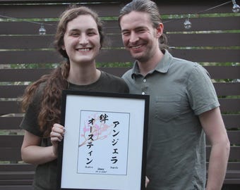 May include: A framed artwork featuring Japanese characters and floral illustrations. The artwork includes the names "Austin" and "Angela" along with a date. The frame is black, and the artwork is held by a person.