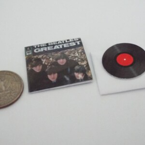 Może przedstawiać: Miniaturowa replika albumu płytowego Beatles&oacute;w z tytułem "The Beatles Greatest" i czarnym winylowym krążkiem z czerwoną etykietą.