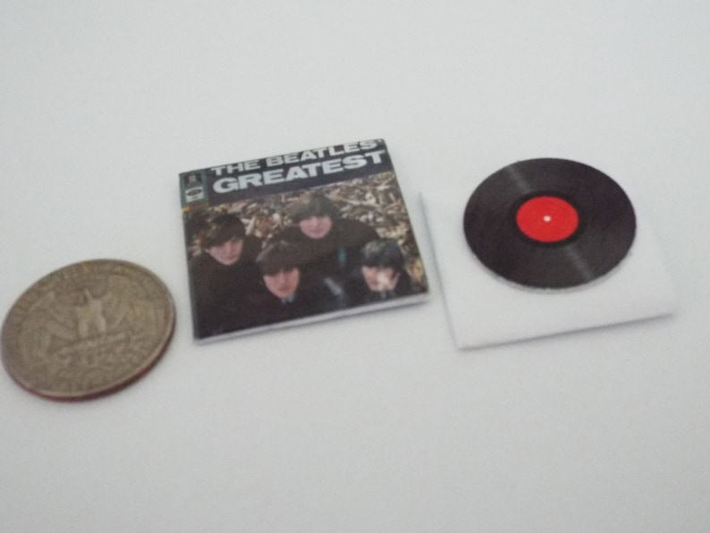 Może przedstawiać: Miniaturowa replika albumu płytowego Beatles&oacute;w z tytułem "The Beatles Greatest" i czarnym winylowym krążkiem z czerwoną etykietą.