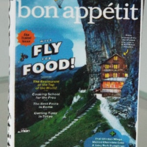 May include: A magazine cover for Bon Appetit featuring a blue sky with a white parachute and the words "WILL FLY FOR FOOD!" in bold, white letters. The background image is a mountainside with a small building at the top of a winding path. The magazine cover also includes text about the restaurant at the top of the world, cooking school for the pros, the best pasta in Rome, and getting tipsy in Tokyo.