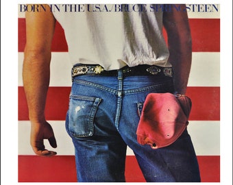 May include: A man wearing a white t-shirt and blue jeans with a red baseball cap in his back pocket. He is standing in front of an American flag.