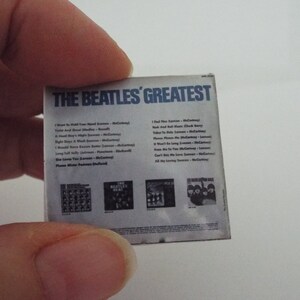 Może przedstawiać: Okładka albumu The Beatles' Greatest przedstawia białe tło z czarnym tekstem i listą utwor&oacute;w. Tytuł albumu jest w kolorze niebieskim. Tylna okładka zawiera cztery mniejsze zdjęcia zespołu.