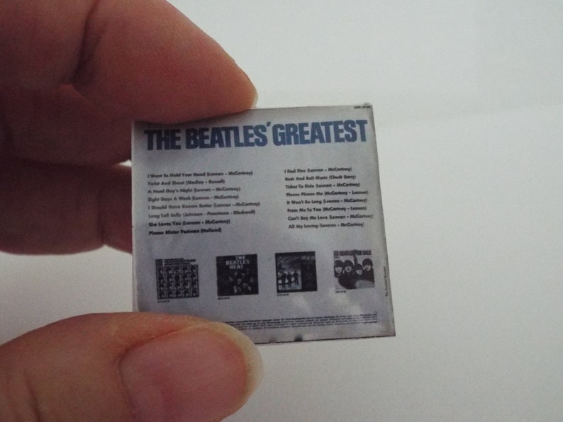 Może przedstawiać: Okładka albumu The Beatles' Greatest przedstawia białe tło z czarnym tekstem i listą utwor&oacute;w. Tytuł albumu jest w kolorze niebieskim. Tylna okładka zawiera cztery mniejsze zdjęcia zespołu.