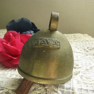 May include: A vintage brass bell with a curved top and a small handle. The word "JAVA" is embossed on the front. A red flower and a dark blue fabric are in the background.