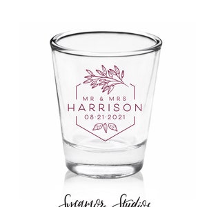May include: A clear glass shot glass with a burgundy design featuring a floral motif, the text "MR & MRS HARRISON 08-21-2021", and a hexagonal frame.