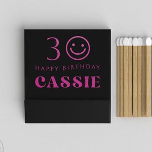 Könnte beinhalten: Eine schwarze Streichholzschachtel mit der Zahl 30 und einem Smiley, den Worten "HAPPY BIRTHDAY" und dem Namen "CASSIE" in Pink. Ein Bündel Holzstreichhölzer mit weißen Spitzen liegt daneben.