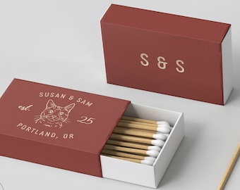 Może przedstawiać: Dwa czerwone pudełka z zapałkami, jedno otwarte, z drewnianymi zapałkami z białymi końcówkami. Otwarte pudełko zawiera napis "SUSAN & SAM" z ilustracją kota i "PORTLAND, OR". Zamknięte pudełko ma napis "S & S" złotymi literami.