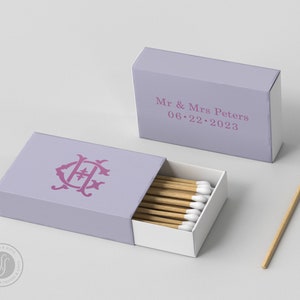 Pode incluir: Duas caixas de fósforos lavanda, uma aberta, mostrando fósforos de madeira com pontas brancas. As caixas apresentam um monograma e o texto "Mr & Mrs Peters 06.22.2023".