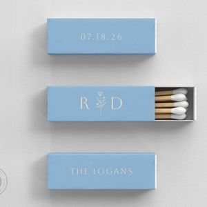 May include: Three light blue matchboxes with gold text. One box displays the date "07.18.26", another has the initials "R D" with a floral design, and the third reads "THE LOGANS". The matchbox in the middle is open, revealing wooden matches.