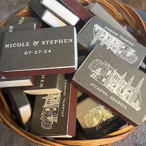 Può includere: Un cesto di vimini pieno di scatole di fiammiferi personalizzate. Le scatole sono grigio scuro con bordi bordeaux. Alcune scatole riportano i nomi "NICOLE & STEPHEN" e la data "07.27.24". Altre presentano una grafica di un edificio e il testo "ATLANTA, GEORGIA".