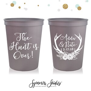 May include: Two gray plastic cups with white text. One cup says "The Hunt is Over!" and the other cup says "Anna & Nate 3.17.18" with a white antler design and floral accents.