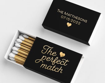 Puede incluir: Cajas de cerillas negras con letras doradas. Una caja está abierta, revelando cerillas con puntas blancas. El texto en las cajas dice "The perfect match" y "THE MATTHESONS 07.15.2022" con un símbolo de corazón.