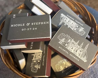 Puede incluir: Una cesta de mimbre llena de cajas de cerillas personalizadas. Las cajas son de color gris oscuro con bordes burdeos. Algunas cajas tienen los nombres "NICOLE & STEPHEN" y la fecha "07.27.24" impresos. Otras cajas tienen un gráfico de un edificio y el texto "ATLANTA, GEORGIA".