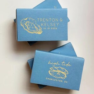 May include: Several light blue matchboxes with gold foil oyster shell designs and text. One box reads "Trenton & Kelsey 10.19.2024". Another reads "high tide" and "Charleston, SC".