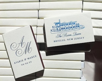 May include: Ivory matchboxes with navy blue text and illustrations. One box features the initials "A M" with the names "Avarie & Marek" and the date "06-06-25". Another box displays a boat illustration and the text "The River Queen, Brielle, New Jersey."
