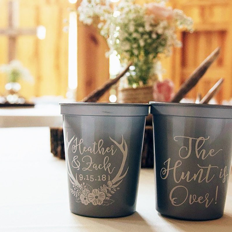 May include: Two gray plastic cups with white text. The first cup says "Heather & Zack 9.15.18" with a floral design and antlers. The second cup says "The Hunt is Over!"