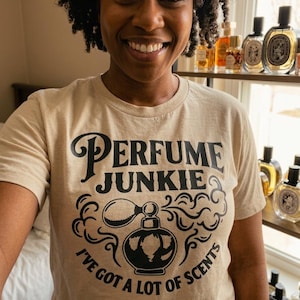 May include: A beige t-shirt with the words "PERFUME JUNKIE" in a stylized font, along with the phrase "I'VE GOT A LOT OF SCENTS" and a perfume bottle graphic. The image also shows a smiling person and a shelf of perfume bottles.
